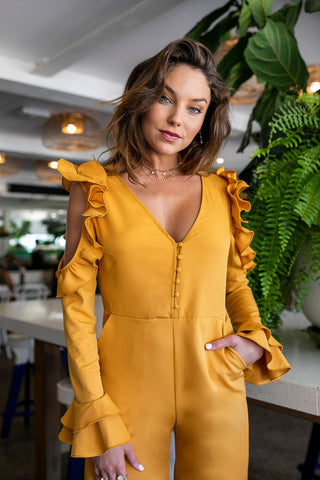 Close up view of the mustard jumpsuit with ruffle cut out long sleeves, button detail and size pockets.