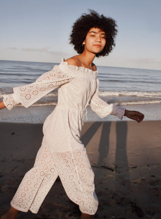 Model on the beach wearing long sleeve off shoulder jumpsuit in beautiful eyelet fabric.