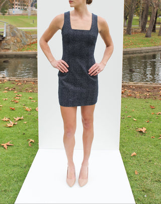 Woman wearing a dark gray dress standing in front of a white backdrop with a park setting in the background.