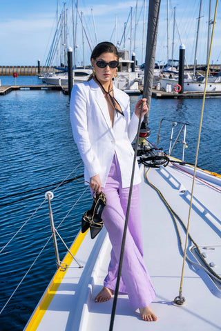 Model wearing white blazer and lilac pant on boat elevated with black heel, black sunglasses and necktie.