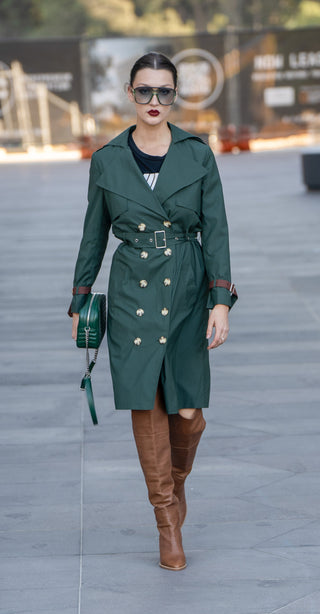 Model wearing green trench coat styled with knee high brown boots.