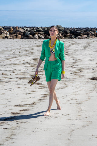 Model walking on the beach wearing matching short and blazer set with yellow bathers and yellow accessories.